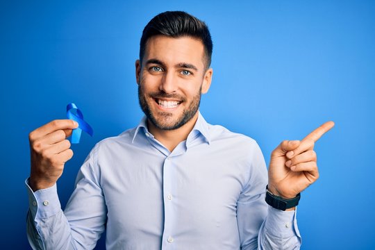 Young handsome man holding blue ribbon as prostate campaing support over blue background very happy pointing with hand and finger to the side