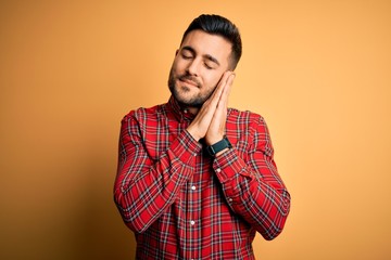 Young handsome man wearing casual shirt standing over isolated yellow background sleeping tired dreaming and posing with hands together while smiling with closed eyes.