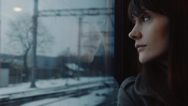 Close-up portrait of beautiful young sad thoughtful Caucasian woman looking out of moving train window slow motion.