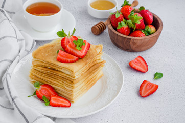 Crepes, traditional Russian thin pancakes on a white plate with fresh strawberries and honey on a light background. Russian maslenitsa. Horizontal orientation. Close up.