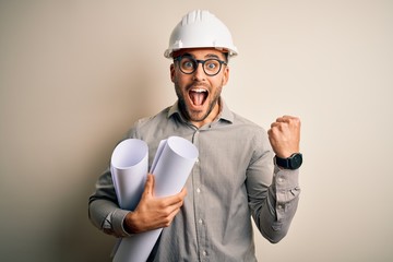 Young architect man wearing contractor helmet holding project paper plan over isolated background...