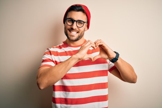 Young handsome man wearing casual striped t-shirt and glasses over white background smiling in love showing heart symbol and shape with hands. Romantic concept.