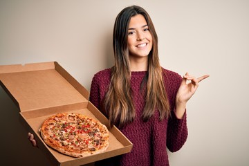 Young beautiful girl holding delivery box with Italian pizza standing over white background very happy pointing with hand and finger to the side