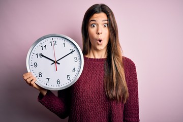 Young beautiful girl doing countdown holding big clock over isolated pink background scared in...