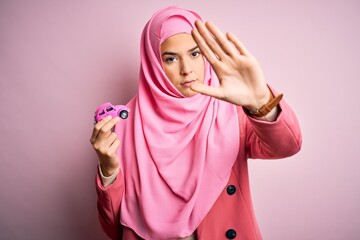 Young girl wearing muslim hijab holding small car toy over isolated pink background with open hand...