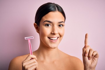 Young beautiful girl using shaver for depilation standing over isolated pink background surprised with an idea or question pointing finger with happy face, number one