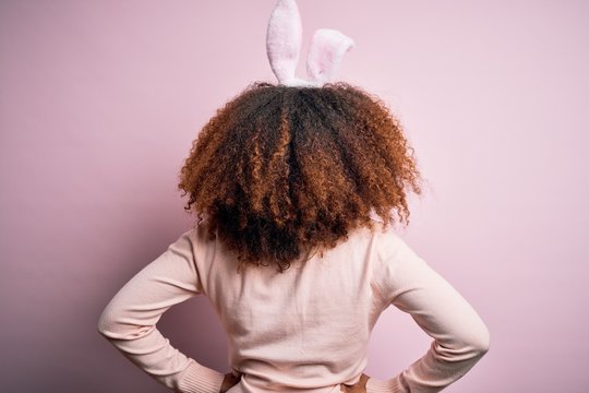 Young african american woman with afro hair wearing bunny ears over pink background standing backwards looking away with arms on body