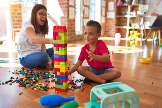 Young beautiful teacher and toddler playing with building blocks around lots of toys at kindergarten