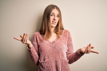Young beautiful redhead woman wearing pink casual sweater over isolated white background clueless...