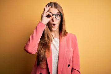 Young beautiful redhead woman wearing jacket and glasses over isolated yellow background doing ok gesture shocked with surprised face, eye looking through fingers. Unbelieving expression.