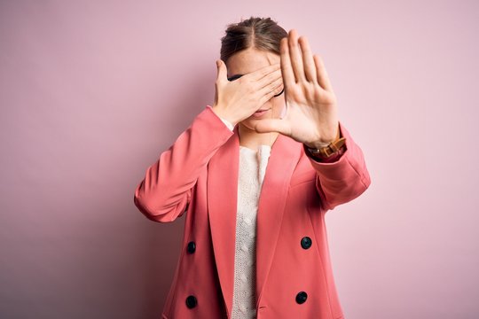 Young beautiful redhead woman wearing jacket and glasses over isolated pink background covering eyes with hands and doing stop gesture with sad and fear expression. Embarrassed and negative concept.