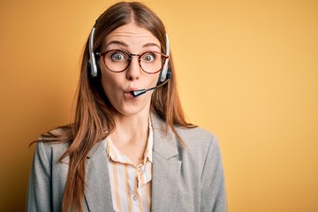 Young redhead call center agent woman overworked wearing glasses using headset afraid and shocked...