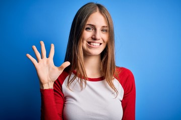 Young beautiful redhead woman wearing casual t-shirt over isolated blue background showing and pointing up with fingers number five while smiling confident and happy.