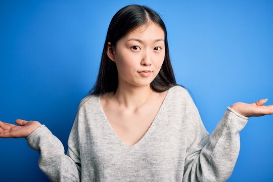 Young beautiful asian woman wearing casual sweater standing over blue isolated background clueless and confused expression with arms and hands raised. Doubt concept.