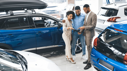 Couple Visiting Car Dealership Talking With Salesman In Auto Showroom