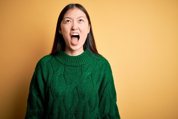 Fototapeta premium Young beautiful asian woman wearing green winter sweater over yellow isolated background angry and mad screaming frustrated and furious, shouting with anger. Rage and aggressive concept.