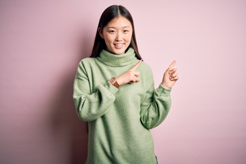 Young beautiful asian woman wearing green winter sweater over pink solated background smiling and looking at the camera pointing with two hands and fingers to the side.