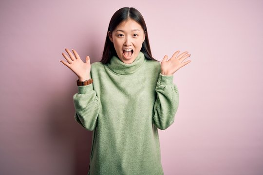 Young beautiful asian woman wearing green winter sweater over pink solated background celebrating crazy and amazed for success with arms raised and open eyes screaming excited. Winner concept