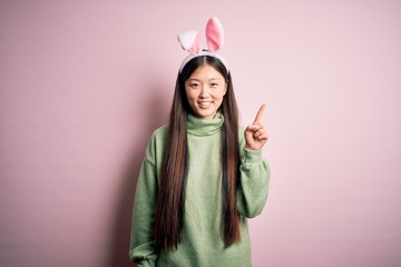 Young asian woman wearing cute easter bunny ears over pink background showing and pointing up with finger number one while smiling confident and happy.