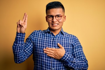 Young handsome latin man wearing casual shirt and glasses over yellow background smiling swearing with hand on chest and fingers up, making a loyalty promise oath