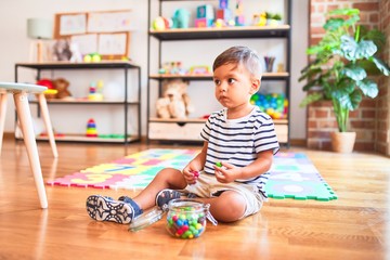 Fototapeta premium Beautiful toddler boy sitting on puzzle eating small colored chocolate balls at kindergarten