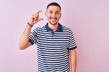 Fototapeta premium Young handsome man wearing nautical striped t-shirt over pink isolated background smiling and confident gesturing with hand doing small size sign with fingers looking and the camera. Measure concept.