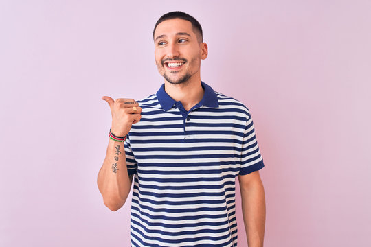 Young handsome man wearing nautical striped t-shirt over pink isolated background smiling with happy face looking and pointing to the side with thumb up.