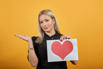 Young blonde girl in black dress on yellow background happy woman holding a piece of paper with a red heart and a hand gesture