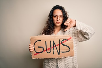 Young beautiful woman with curly hair holding banner with prohibited guns message with angry face,...