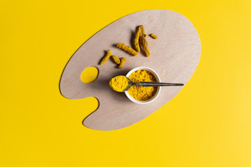 Turmeric spice powder in bowl with spoon and roots on wooden palette board on yellow  background with copy space