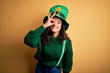 Beautiful curly hair woman wearing green hat with clover celebrating saint patricks day doing ok gesture with hand smiling, eye looking through fingers with happy face.