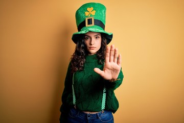 Beautiful curly hair woman wearing green hat with clover celebrating saint patricks day doing stop sing with palm of the hand. Warning expression with negative and serious gesture on the face.