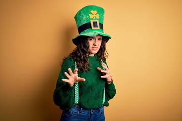Beautiful curly hair woman wearing green hat with clover celebrating saint patricks day disgusted expression, displeased and fearful doing disgust face because aversion reaction. With hands raised