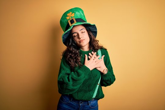 Beautiful curly hair woman wearing green hat with clover celebrating saint patricks day smiling with hands on chest with closed eyes and grateful gesture on face. Health concept.