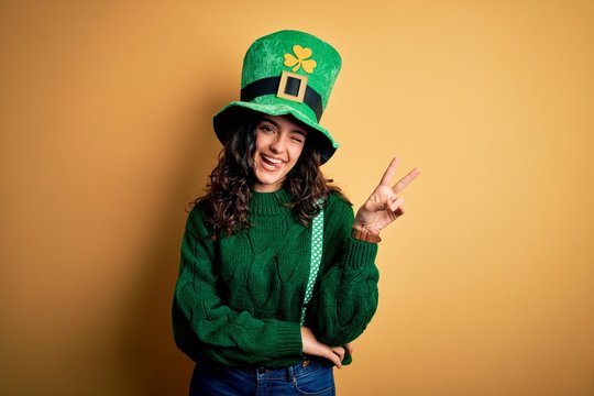 Beautiful curly hair woman wearing green hat with clover celebrating saint patricks day smiling with happy face winking at the camera doing victory sign. Number two.