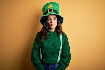 Beautiful curly hair woman wearing green hat with clover celebrating saint patricks day making fish face with lips, crazy and comical gesture. Funny expression.