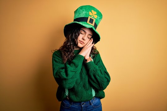 Beautiful curly hair woman wearing green hat with clover celebrating saint patricks day sleeping tired dreaming and posing with hands together while smiling with closed eyes.