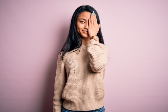 Young beautiful chinese woman wearing casual sweater over isolated pink background covering one eye with hand, confident smile on face and surprise emotion.