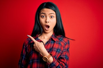 Young beautiful chinese woman wearing casual shirt over isolated red background Surprised pointing with finger to the side, open mouth amazed expression.