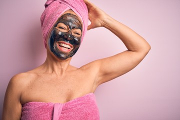 Middle age brunette woman wearing beauty black face mask over isolated pink background smiling confident touching hair with hand up gesture, posing attractive and fashionable