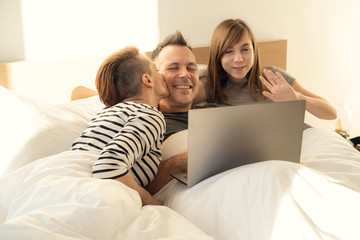 Happy relaxed family of using laptop in bed at home. Mom, father and daughter spending time together in the morning