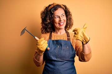 Middle age gardener woman wearing apron holding rake over isolated yellow background pointing and...