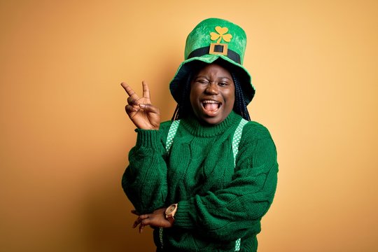 Plus size african american woman with braids wearing green hat with clover on st patricks day smiling with happy face winking at the camera doing victory sign. Number two.