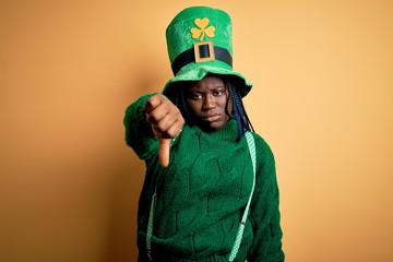 Plus size african american woman with braids wearing green hat with clover on st patricks day looking unhappy and angry showing rejection and negative with thumbs down gesture. Bad expression.