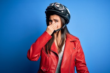 Young beautiful brunette motorcycliste woman wearing motorcycle helmet and jacket smelling something stinky and disgusting, intolerable smell, holding breath with fingers on nose. Bad smell