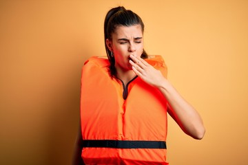 Young beautiful brunette woman wearing orange safe lifejacket over yellow background bored yawning...