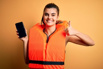 Young beautiful brunette woman wearing safe lifejacket showing smartphone happy with big smile...