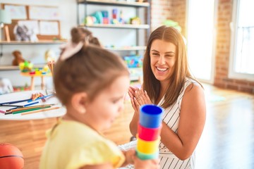 Young beautiful teacher and toddler playing with plastic cups toy at kindergarten