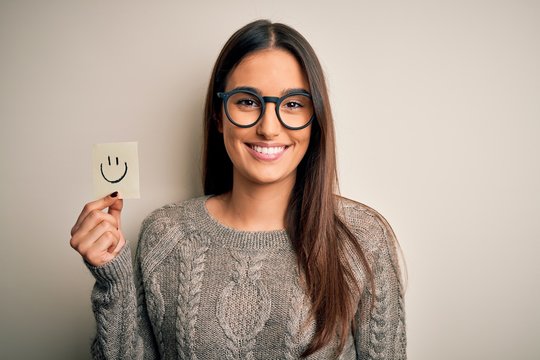 Young beautiful brunette woman wearing glasses holding paper with smile emoji with a happy face standing and smiling with a confident smile showing teeth