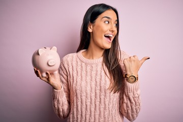 Young beautiful brunette woman holding piggy bank saving money for retirement pointing and showing...